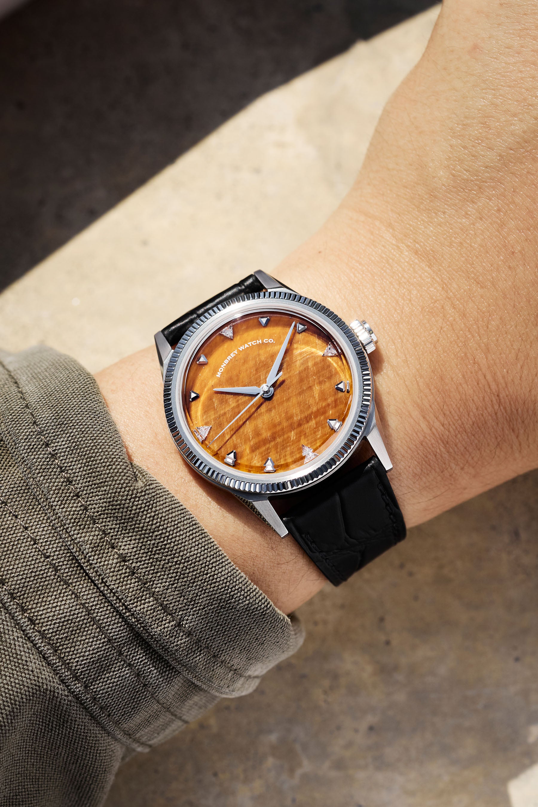 A wristwatch with a round silver-tone case, with a silver-tone fluted bezel, and a natural tiger eye stone dial, and a black croco leather strap, worn by a person in a casual setting. Lab-grown diamond on dial.
