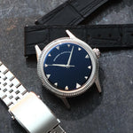 A wristwatch with a round silver-tone case and fluted bezel, a natural Onyx stone dial, and a black croco leather strap, styled on an dark textured surface background next to a silver jubilee bracelet. Lab-grown diamond on dial.