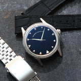 A wristwatch with a round silver-tone case and fluted bezel, a natural Onyx stone dial, and a black croco leather strap, styled on an dark textured surface background next to a silver jubilee bracelet. Lab-grown diamond on dial.