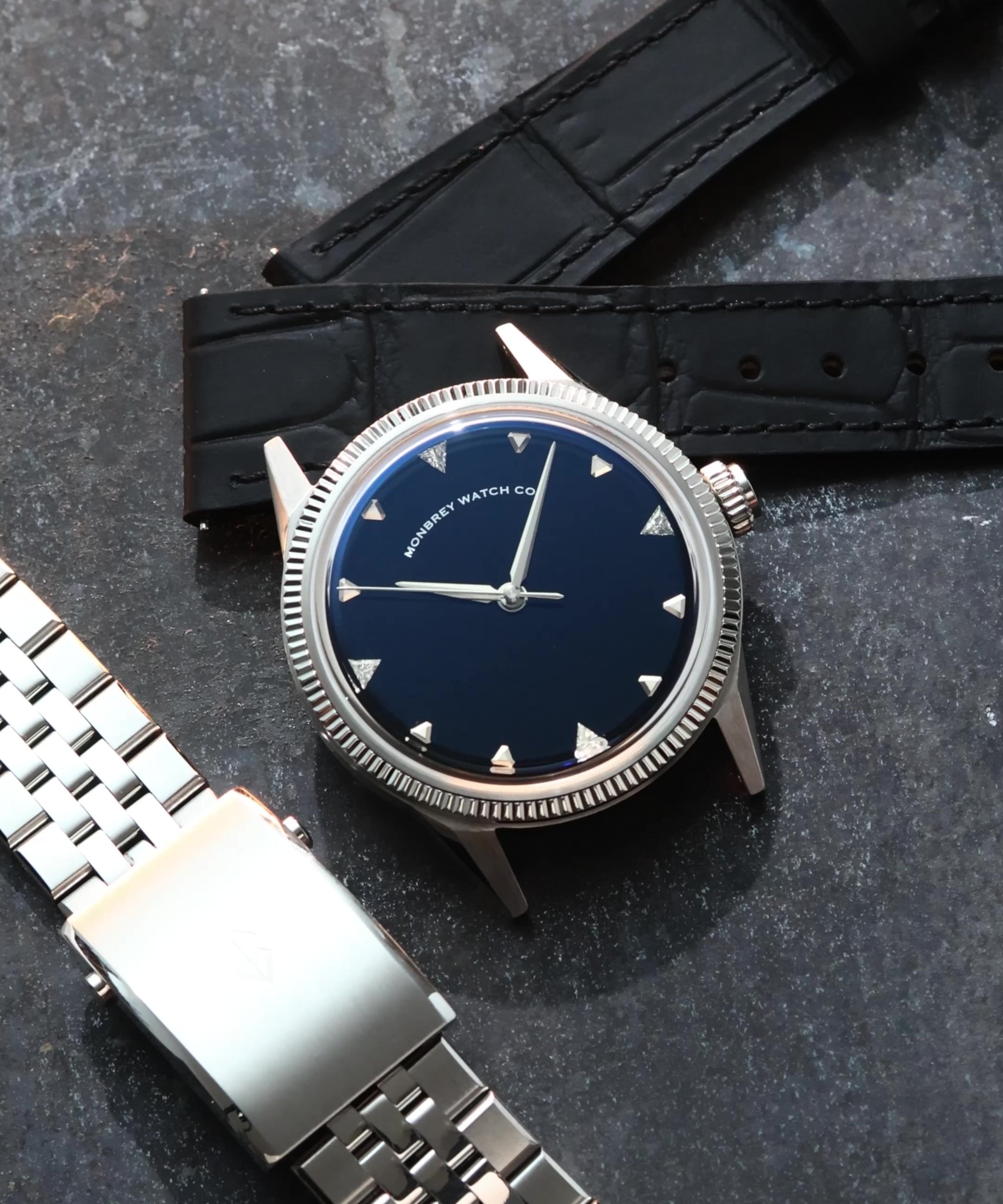 A wristwatch with a round silver-tone case and fluted bezel, a natural Onyx stone dial, and a black croco leather strap, styled on an dark textured surface background next to a silver jubilee bracelet. Lab-grown diamond on dial.