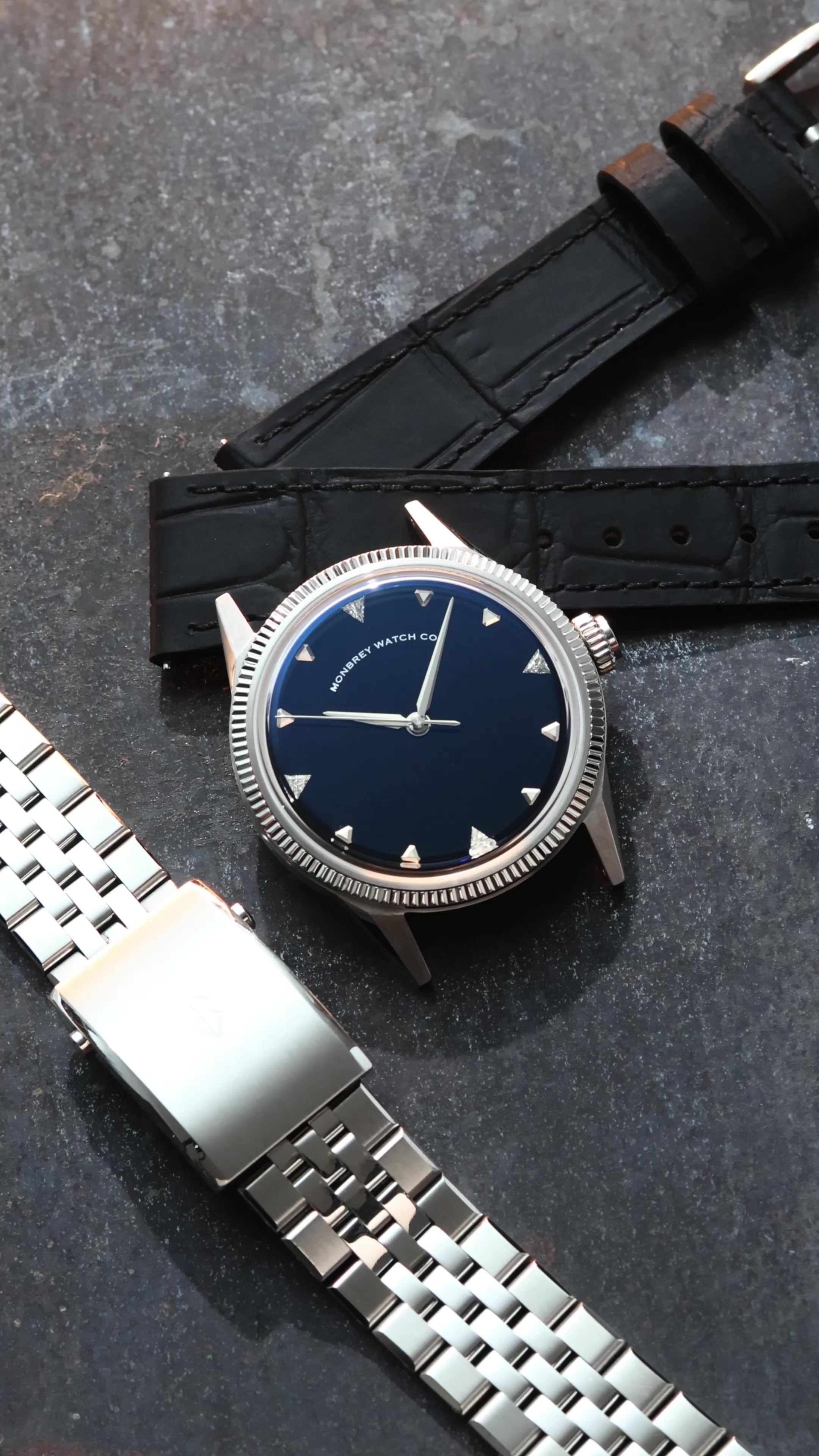 A wristwatch with a round silver-tone case and fluted bezel, a natural Onyx stone dial, and a black croco leather strap, styled on an dark textured surface background next to a silver jubilee bracelet. Lab-grown diamond on dial.