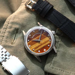 A wristwatch with a round silver-tone case, a natural tiger eye stone dial, and a black croco leather strap, styled on an olive green fabric background next to a silver metal bracelet. Lab-grown diamond on dial.