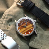 A wristwatch with a round silver-tone case, a natural tiger eye stone dial, and a black croco leather strap, styled on an olive green fabric background next to a silver metal bracelet. Lab-grown diamond on dial.