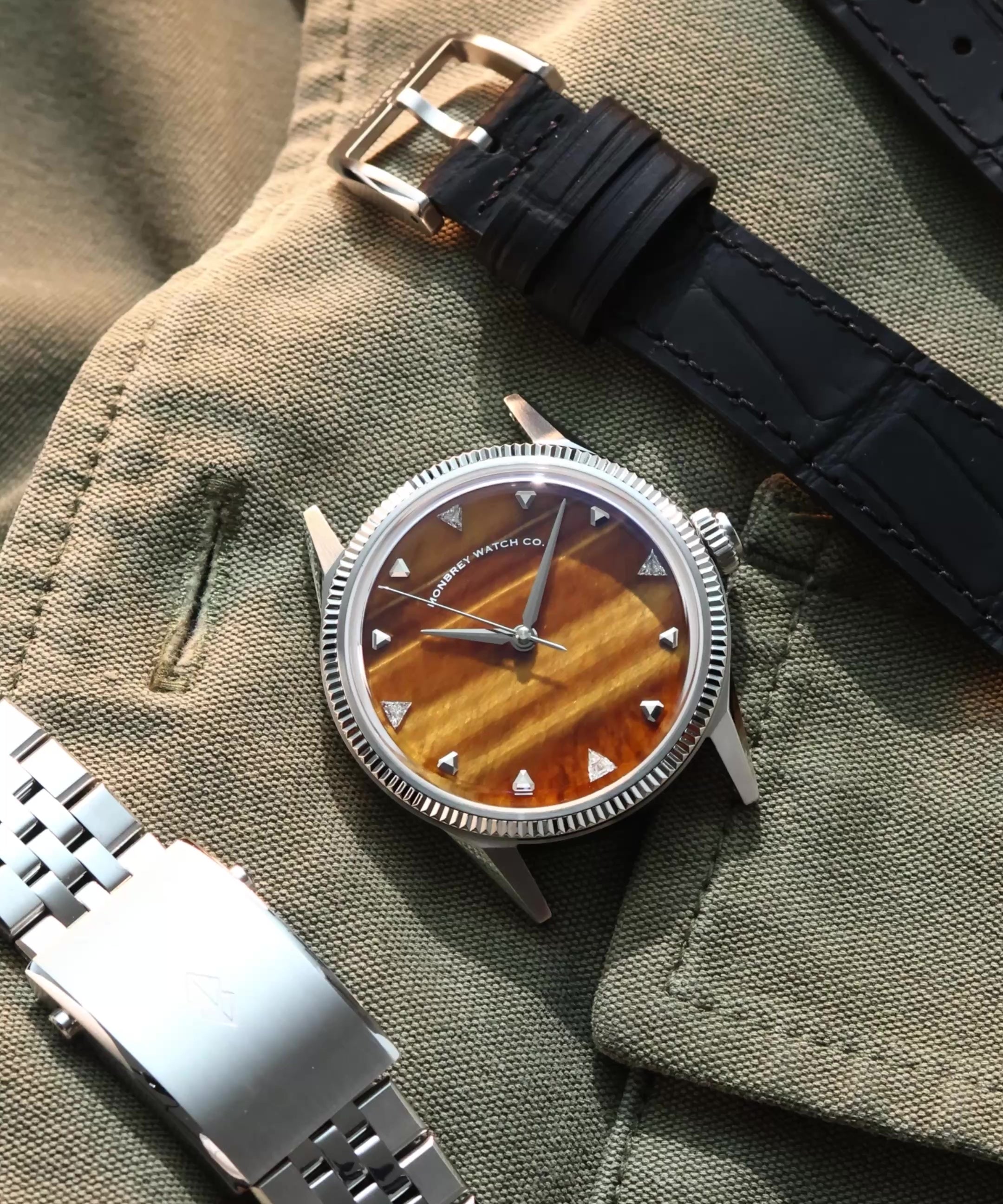 A wristwatch with a round silver-tone case, a natural tiger eye stone dial, and a black croco leather strap, styled on an olive green fabric background next to a silver metal bracelet. Lab-grown diamond on dial.