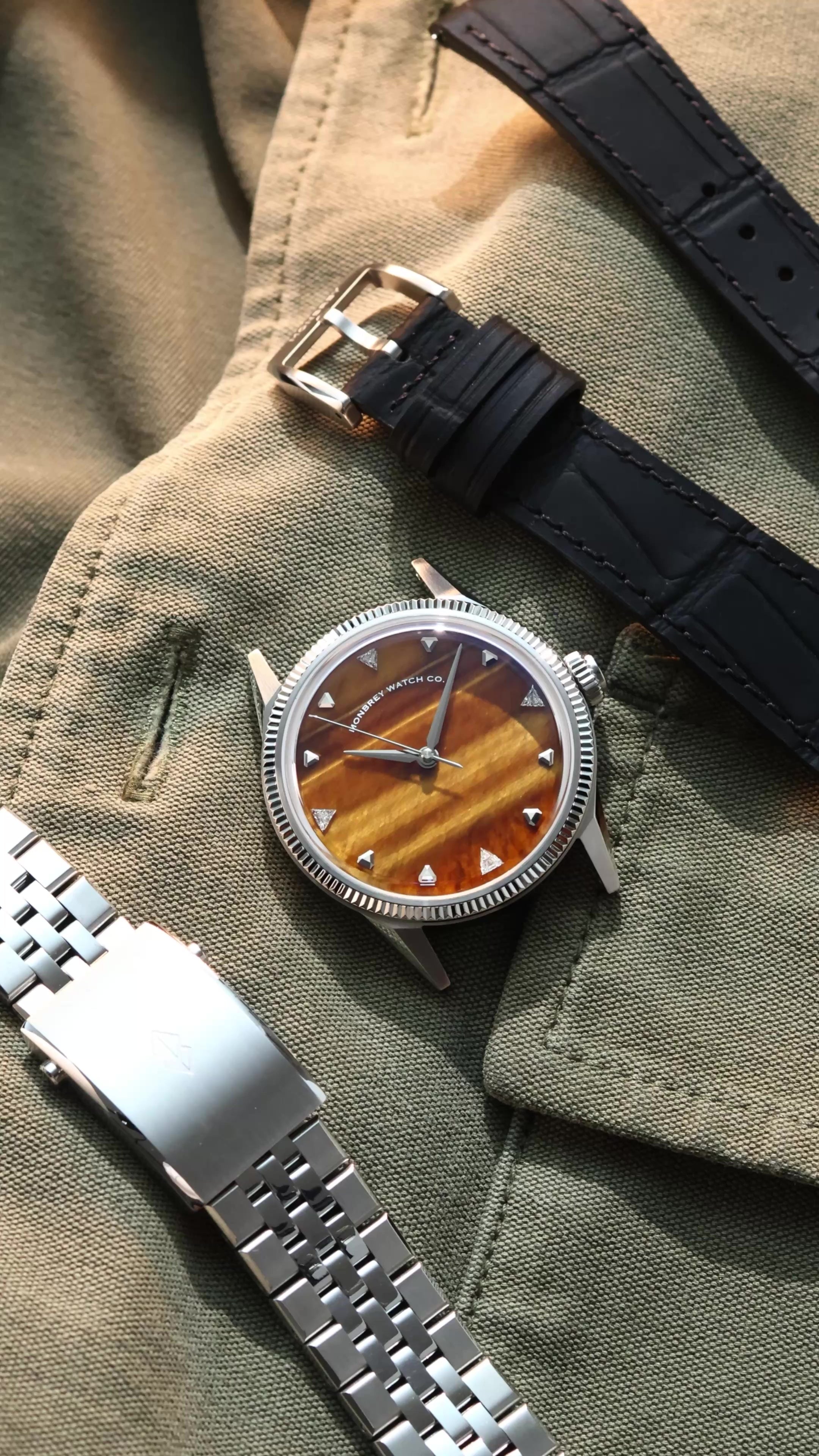 A wristwatch with a round silver-tone case, a natural tiger eye stone dial, and a black croco leather strap, styled on an olive green fabric background next to a silver metal bracelet. Lab-grown diamond on dial.
