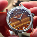 A close-up of a wristwatch with a silver-tone fluted bezel and a natural tiger eye stone dial, held by two hands. Lab-grown diamond on dial.
