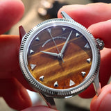 A close-up of a wristwatch with a silver-tone fluted bezel and a natural tiger eye stone dial, held by two hands. Lab-grown diamond on dial.