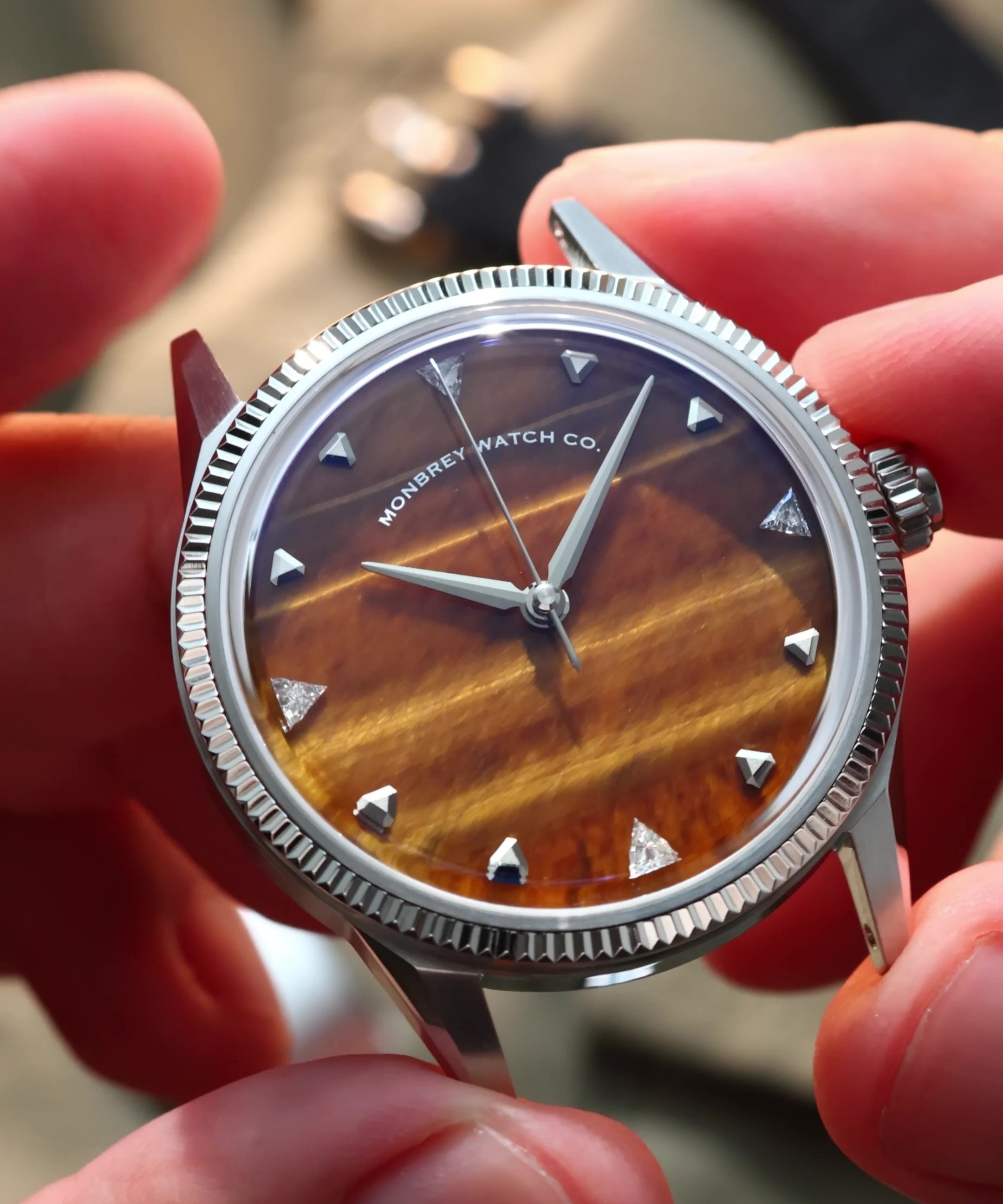 A close-up of a wristwatch with a silver-tone fluted bezel and a natural tiger eye stone dial, held by two hands. Lab-grown diamond on dial.