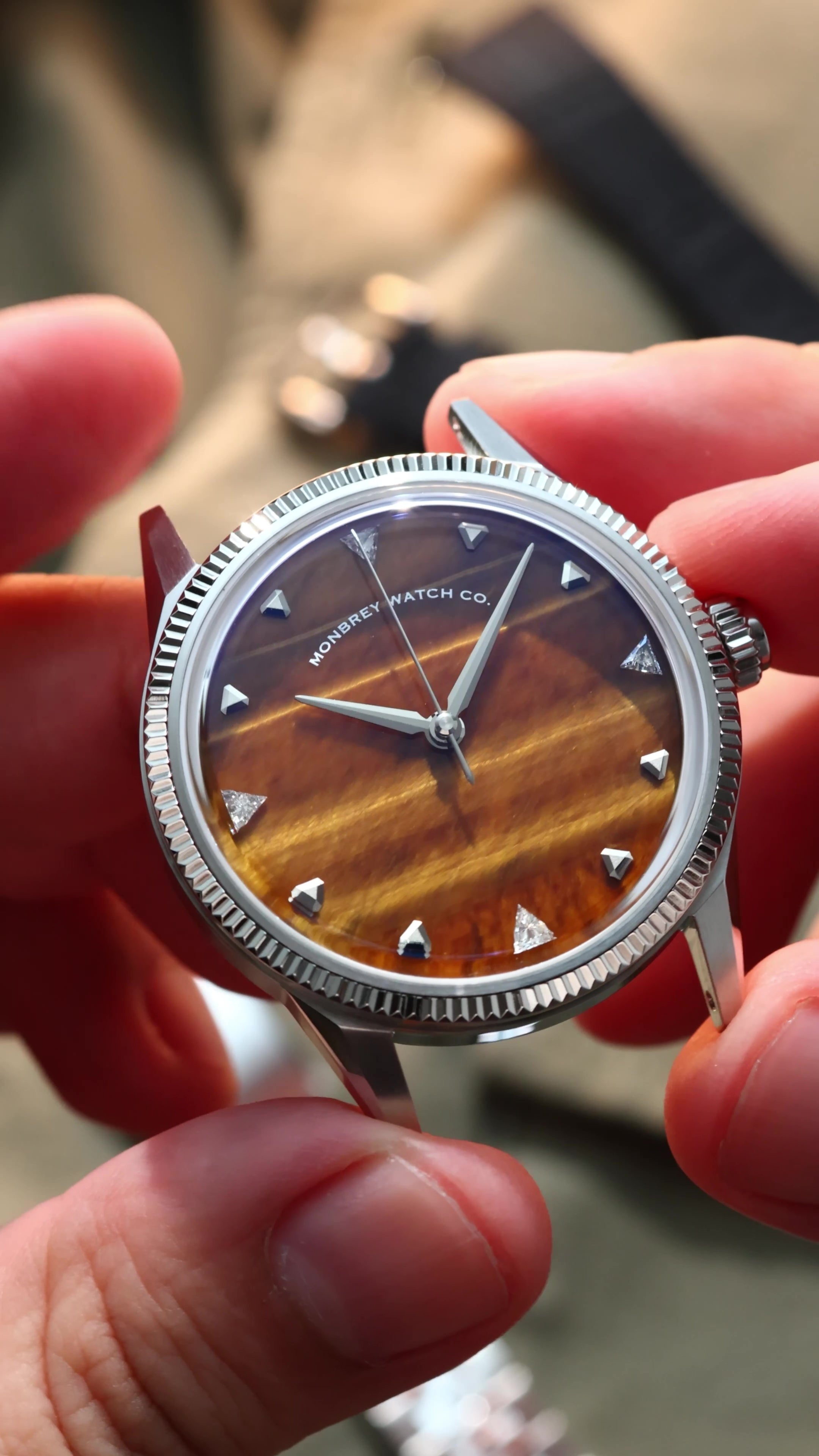 A close-up of a wristwatch with a silver-tone fluted bezel and a natural tiger eye stone dial, held by two hands. Lab-grown diamond on dial.