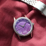 A silver-tone wristwatch with a flusted bezel and a Natural Charoite Stone dial, displayed next to its silver metal bracelet on a draped burgundy fabric background.