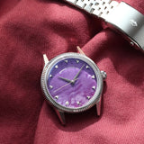 A silver-tone wristwatch with a flusted bezel and a Natural Charoite Stone dial, displayed next to its silver metal bracelet on a draped burgundy fabric background.