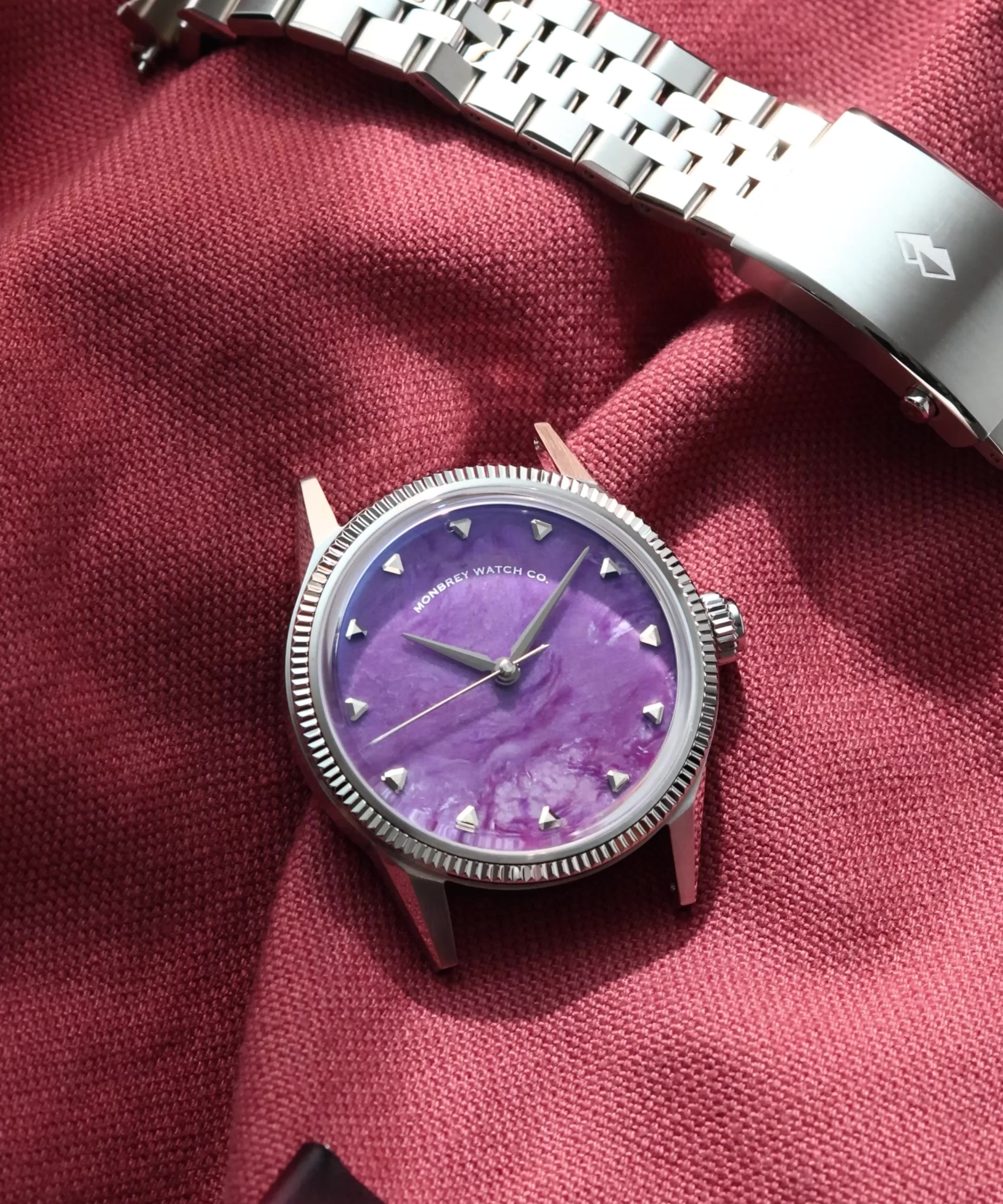 A silver-tone wristwatch with a flusted bezel and a Natural Charoite Stone dial, displayed next to its silver metal bracelet on a draped burgundy fabric background.