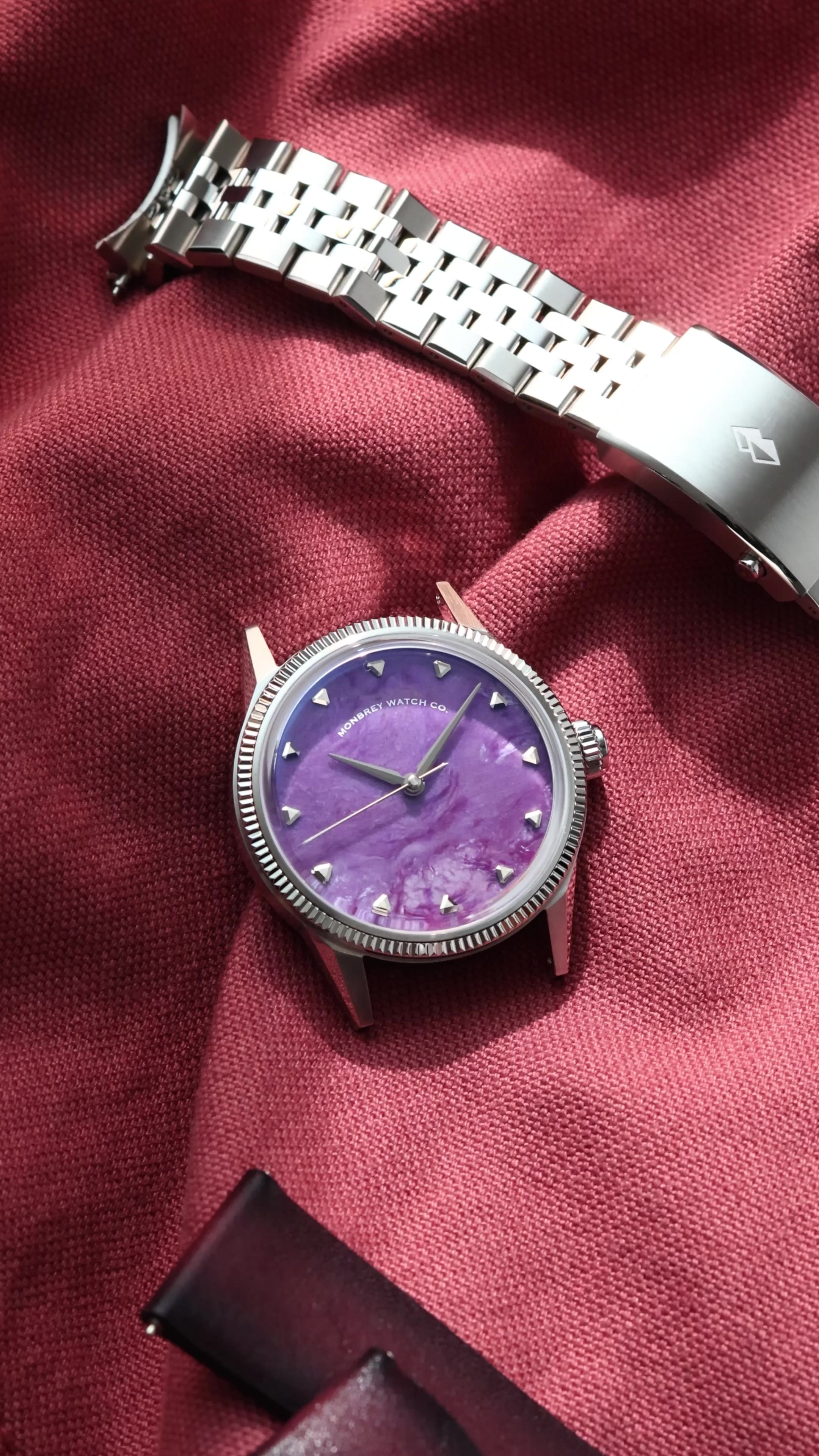 A silver-tone wristwatch with a flusted bezel and a Natural Charoite Stone dial, displayed next to its silver metal bracelet on a draped burgundy fabric background.