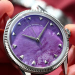 A close-up of a wristwatch with a natural Charoite dial and silver-tone case and fluted, held in a person's hand against a blurred red fabric background. 