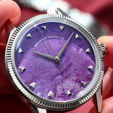 A close-up of a wristwatch with a natural Charoite dial and silver-tone case and fluted, held in a person's hand against a blurred red fabric background. 