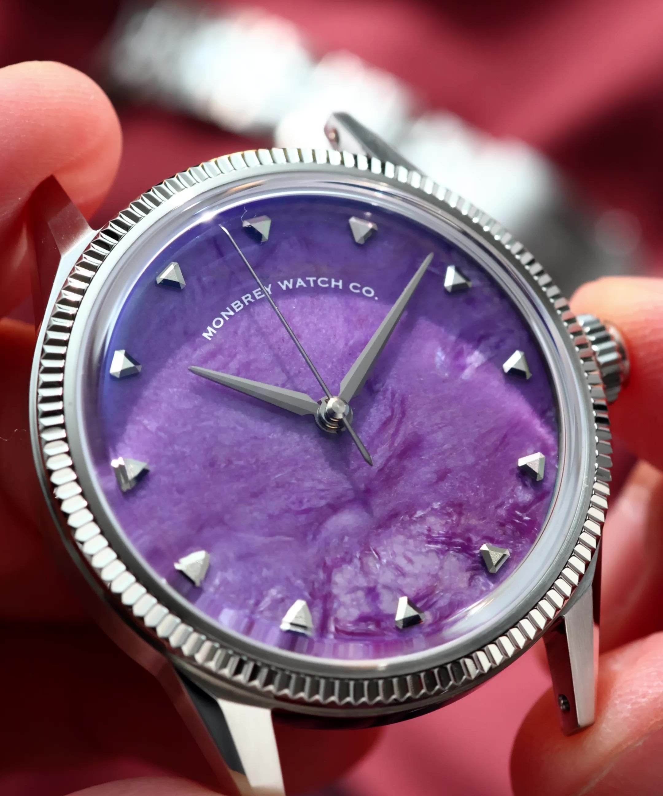 A close-up of a wristwatch with a natural Charoite dial and silver-tone case and fluted, held in a person's hand against a blurred red fabric background. 