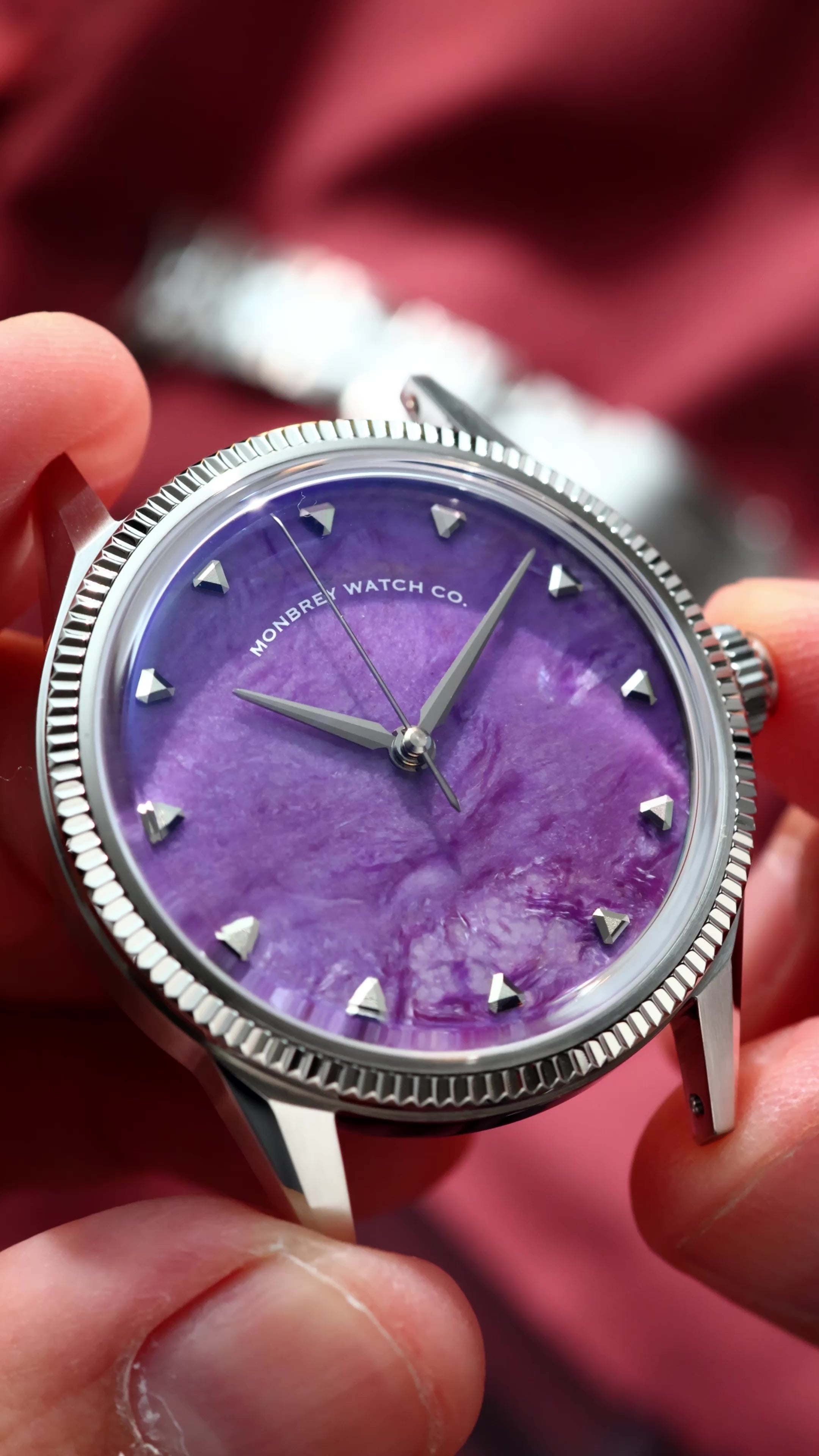 A close-up of a wristwatch with a natural Charoite dial and silver-tone case and fluted, held in a person's hand against a blurred red fabric background. 