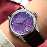 A wristwatch with a natural charoite stone dial and silver-toned case and fluted, worn on a person's wrist against a dark red fabric background.