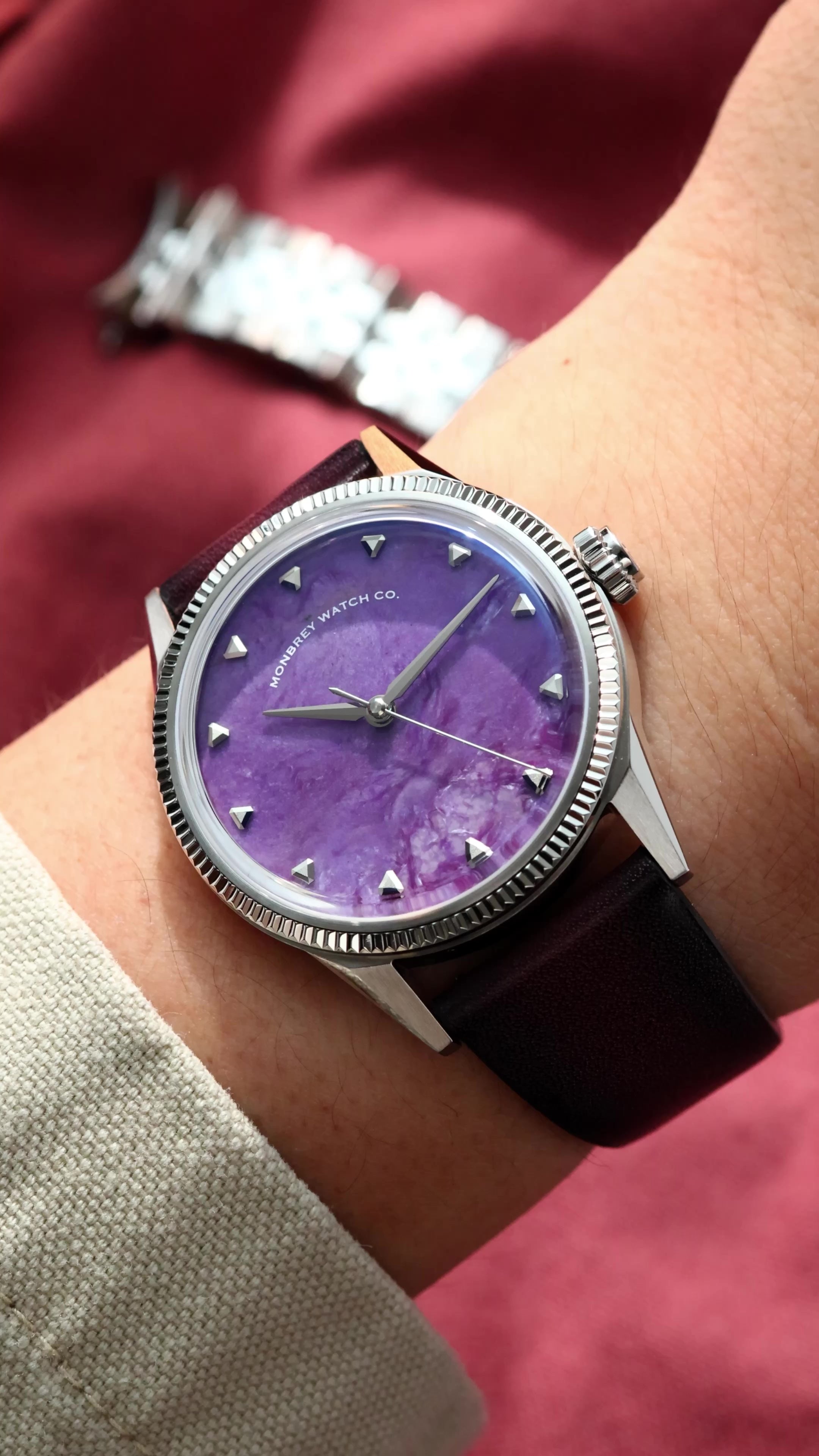 A wristwatch with a natural charoite stone dial and silver-toned case and fluted, worn on a person's wrist against a dark red fabric background.