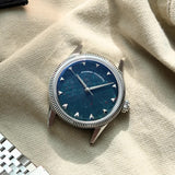 A luxury wristwatch with a natural bloodstone dial and silver-toned case and fluted bezel, styled on a beige canvas background next to a silver jubilee bracelet.