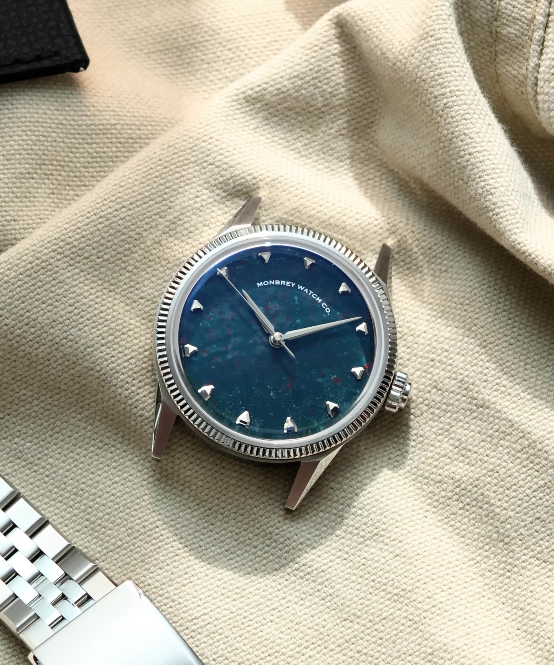 A luxury wristwatch with a natural bloodstone dial and silver-toned case and fluted bezel, styled on a beige canvas background next to a silver jubilee bracelet.