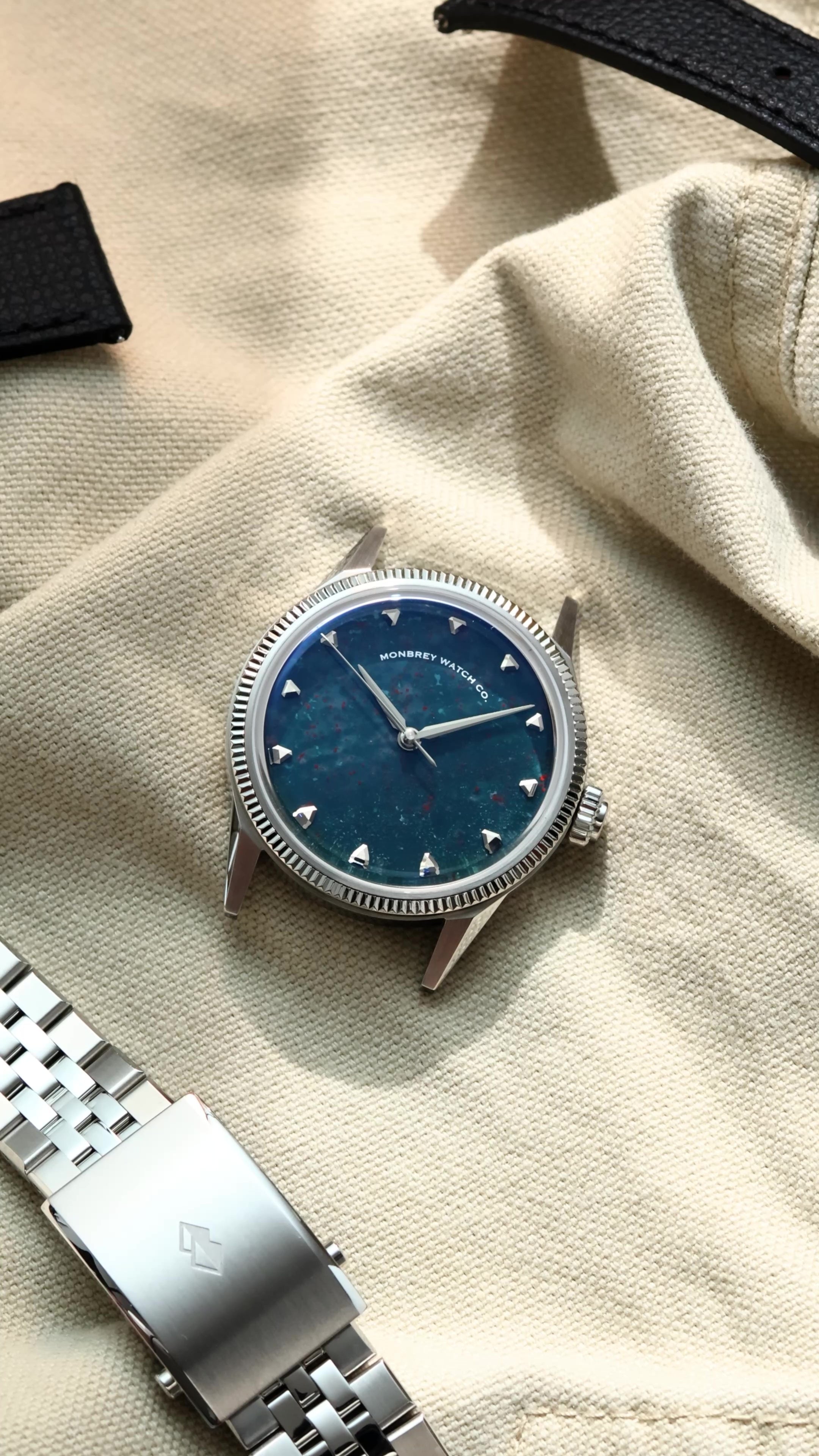 A luxury wristwatch with a natural bloodstone dial and silver-toned case and fluted bezel, styled on a beige canvas background next to a silver jubilee bracelet.