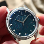 A close-up of a wristwatch being held by hands. The watch has a silver-tone fluted bezel, a natural bloodstone speckled dial, and silver-tone hour markers.