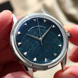 A close-up of a wristwatch being held by hands. The watch has a silver-tone fluted bezel, a natural bloodstone speckled dial, and silver-tone hour markers.