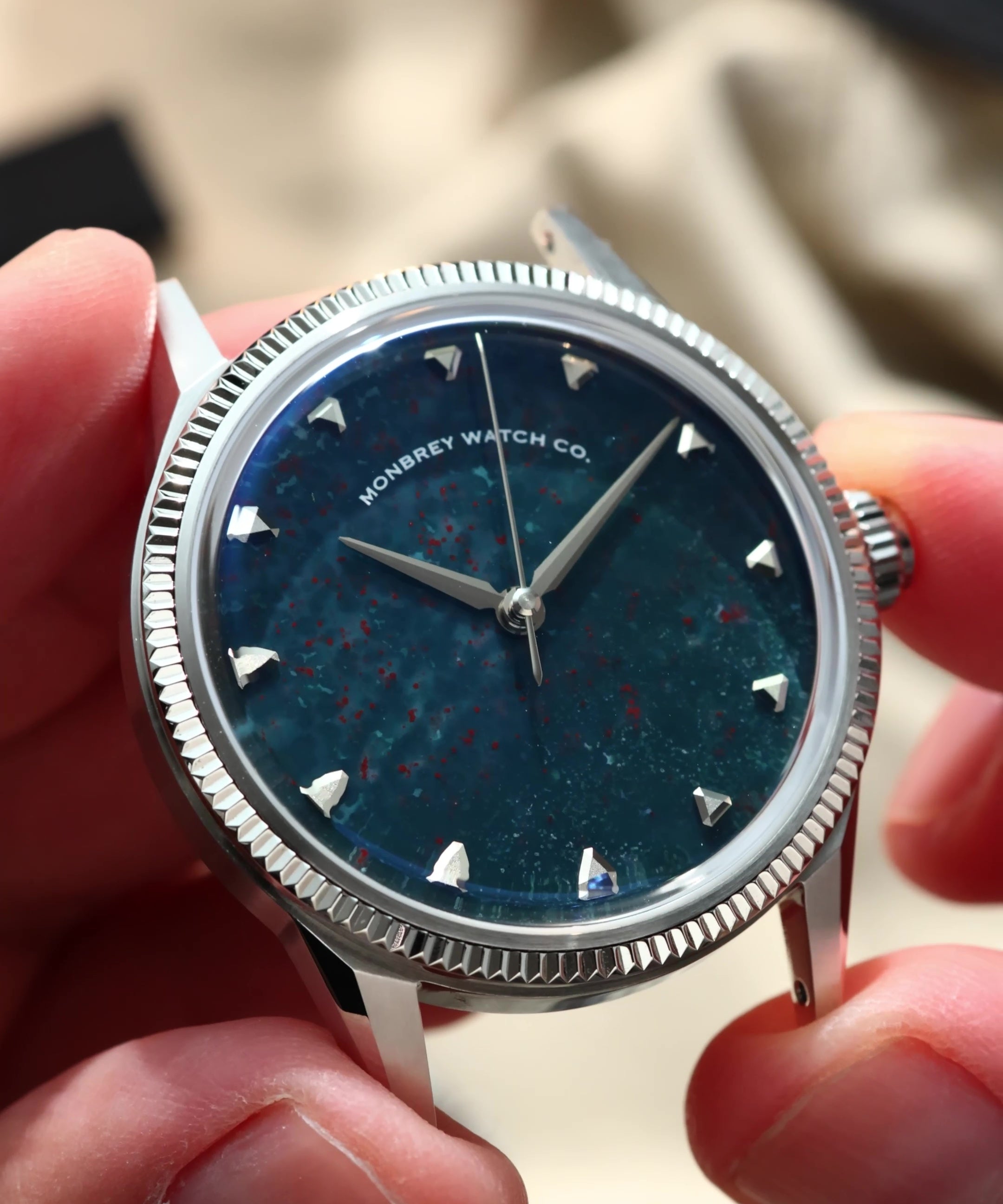 A close-up of a wristwatch being held by hands. The watch has a silver-tone fluted bezel, a natural bloodstone speckled dial, and silver-tone hour markers.