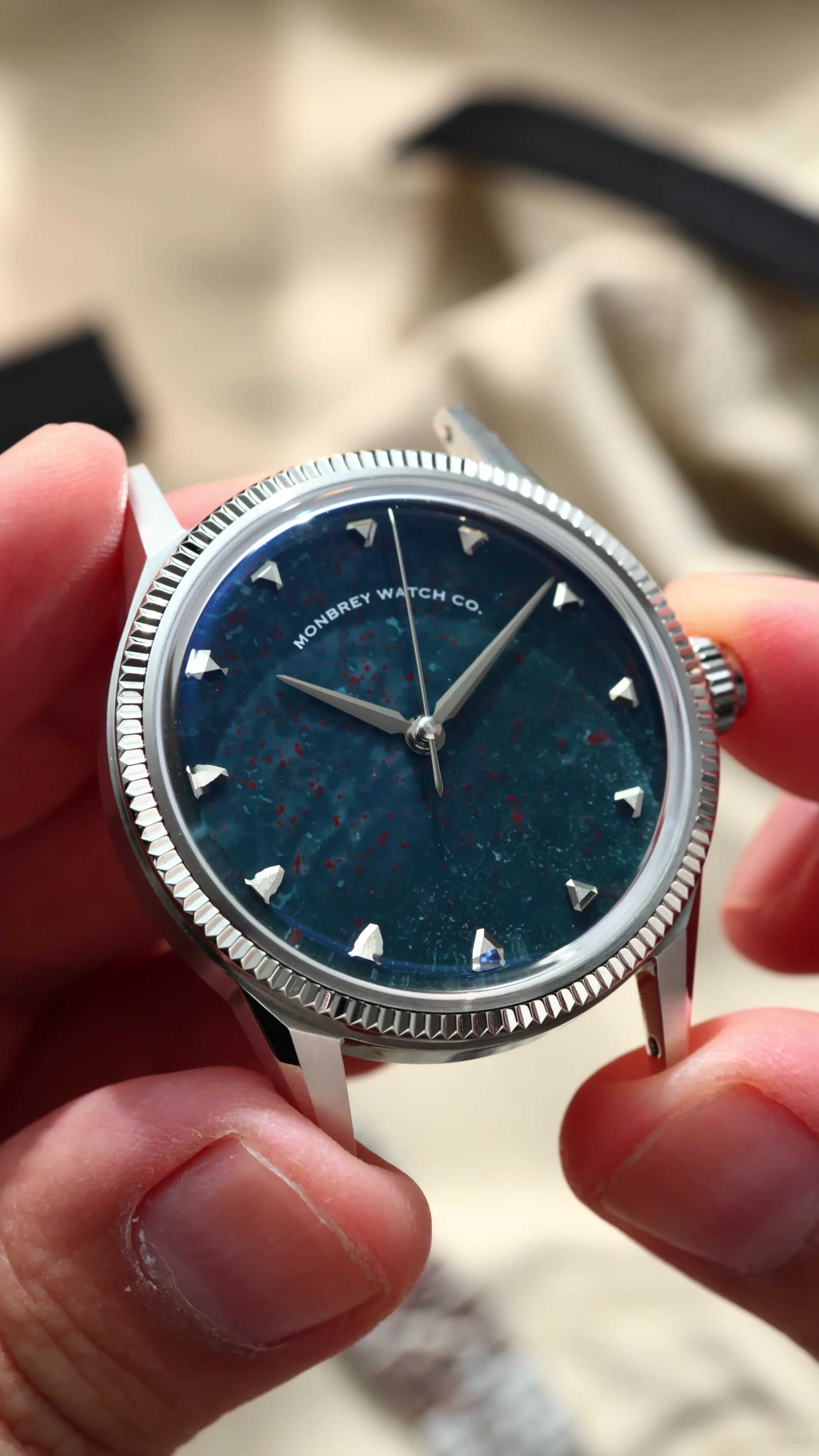 A close-up of a wristwatch being held by hands. The watch has a silver-tone fluted bezel, a natural bloodstone speckled dial, and silver-tone hour markers.