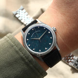 A wristwatch with a natural bloodstone dial, silver-tone case and fluted bezel, and black leather strap, shown worn on a person's wrist against a beige fabric background.