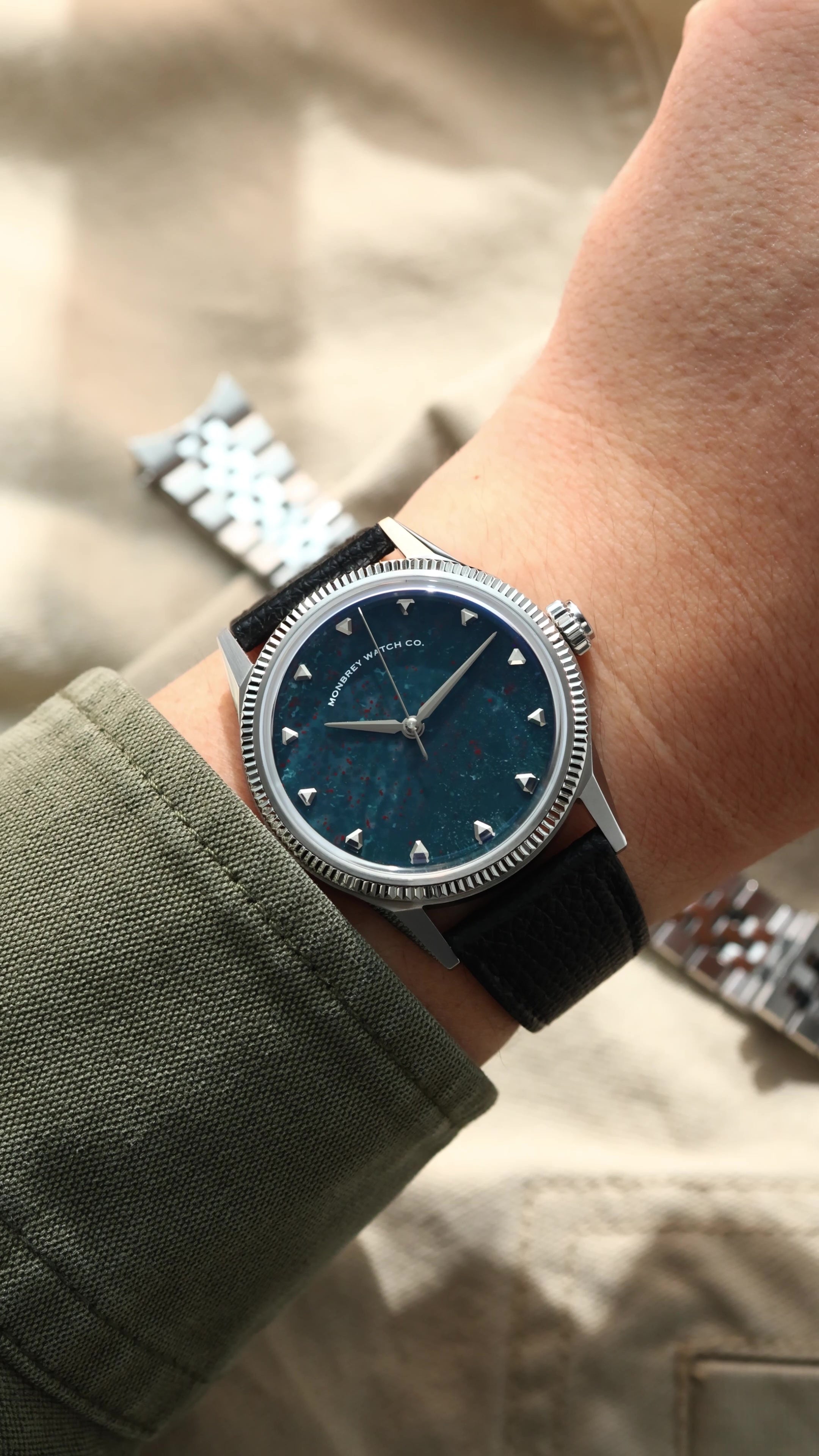A wristwatch with a natural bloodstone dial, silver-tone case and fluted bezel, and black leather strap, shown worn on a person's wrist against a beige fabric background.