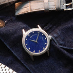 A wristwatch with a natural Sodalite stone dial and silver-toned case, styled on dark navy velvet fabric alongside a gray alligator-embossed strap and a silver jubilee bracelet.