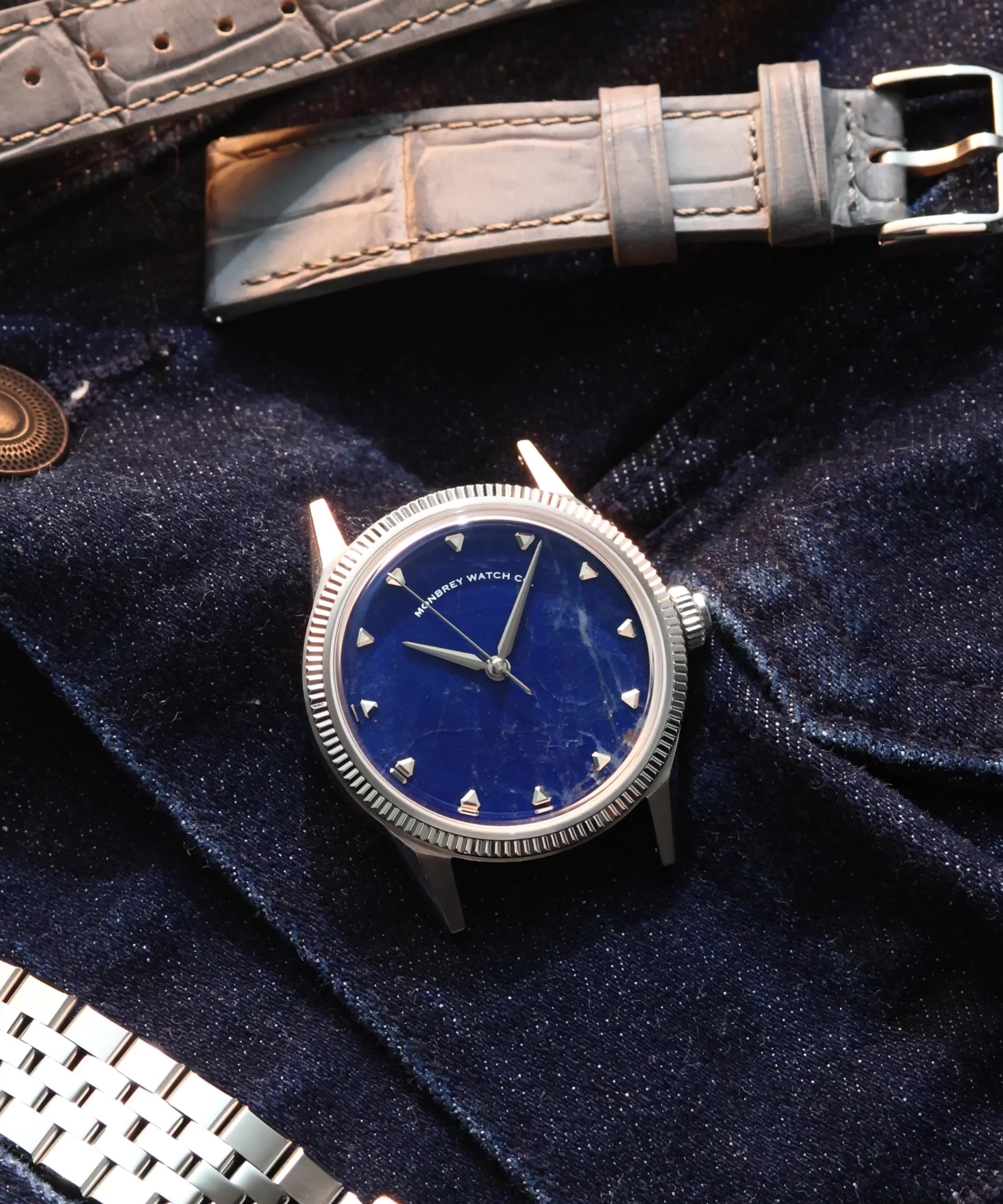 A wristwatch with a natural Sodalite stone dial and silver-toned case, styled on dark navy velvet fabric alongside a gray alligator-embossed strap and a silver jubilee bracelet.