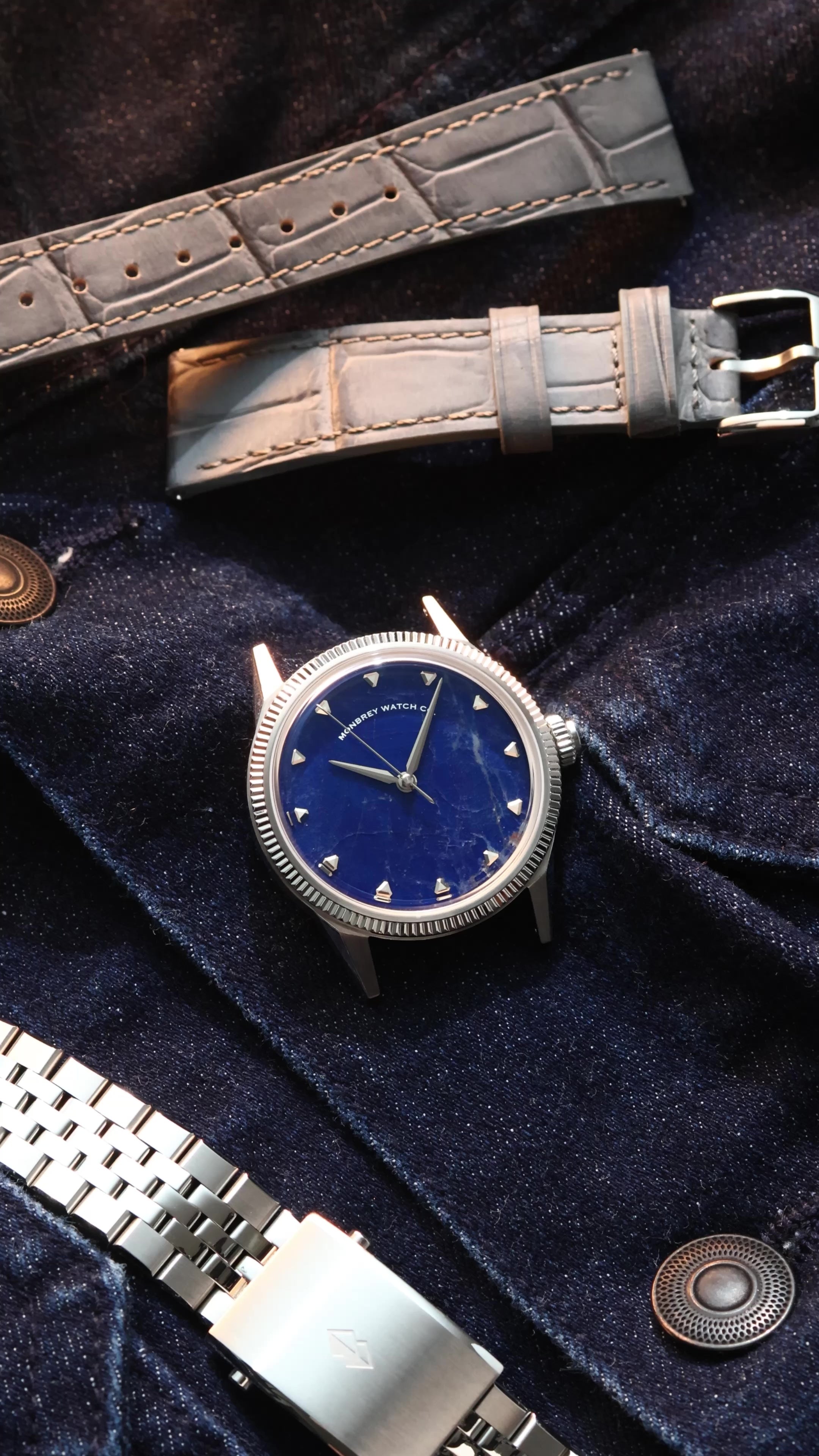 A wristwatch with a natural Sodalite stone dial and silver-toned case, styled on dark navy velvet fabric alongside a gray alligator-embossed strap and a silver jubilee bracelet.