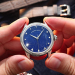 A close-up of a wristwatch with a natural Sodalite stone dial and silver-tone case, held between two hands. The watch has a fluted silver bezel, silver hour markers, and silver hands.