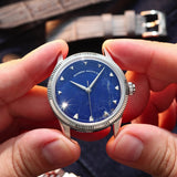 A close-up of a wristwatch with a natural Sodalite stone dial and silver-tone case, held between two hands. The watch has a fluted silver bezel, silver hour markers, and silver hands.
