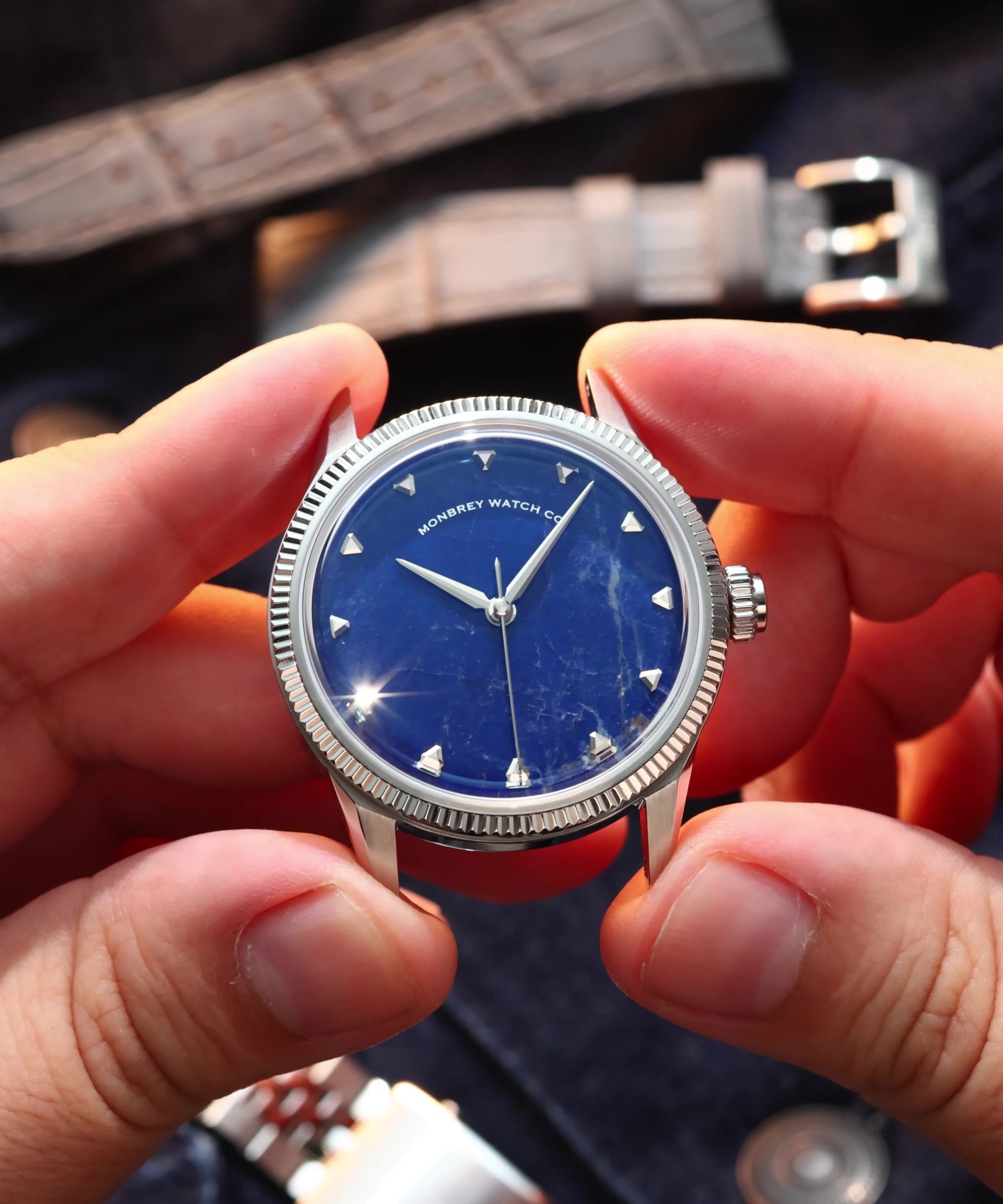 A close-up of a wristwatch with a natural Sodalite stone dial and silver-tone case, held between two hands. The watch has a fluted silver bezel, silver hour markers, and silver hands.
