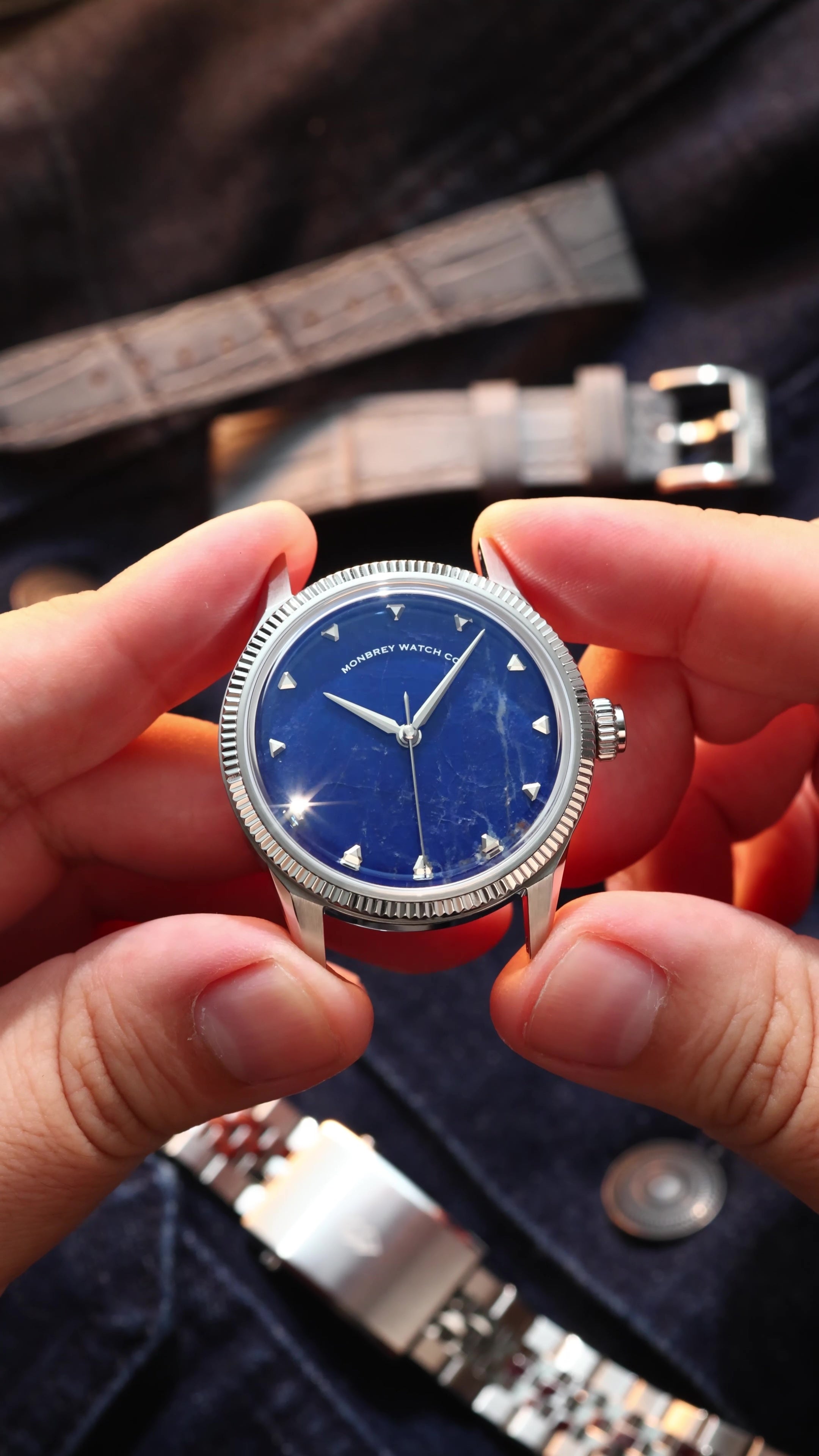 A close-up of a wristwatch with a natural Sodalite stone dial and silver-tone case, held between two hands. The watch has a fluted silver bezel, silver hour markers, and silver hands.