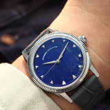 A close-up of a wrist wearing a vintage-style watch with a natural Sodalite stone dial, silver-tone case with fluted bezel, and dark gray leather strap.