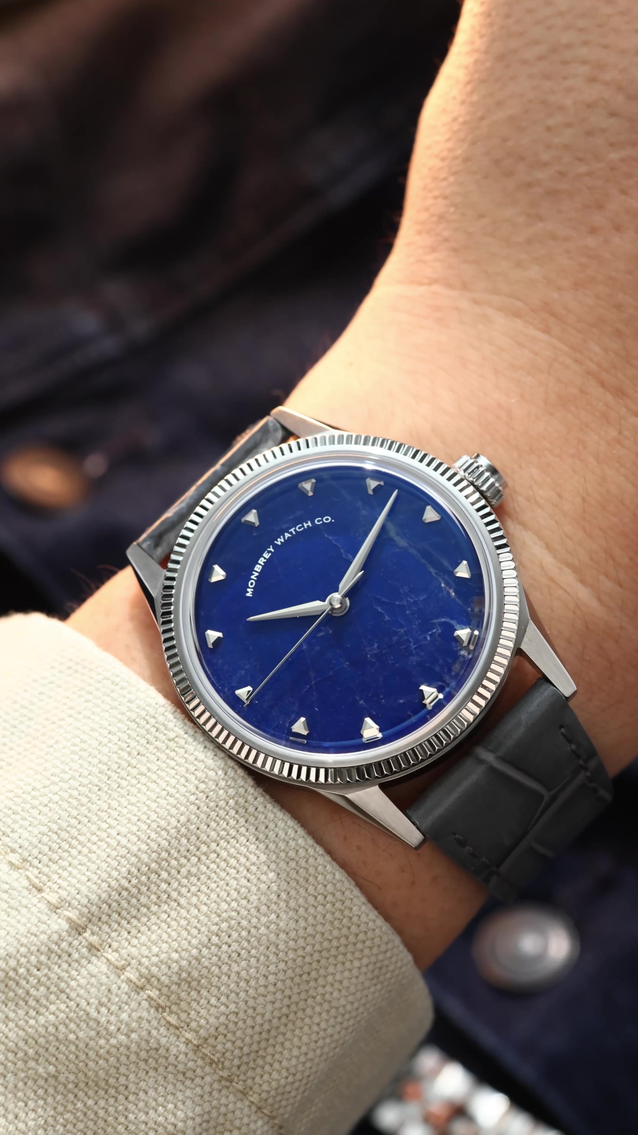 A close-up of a wrist wearing a vintage-style watch with a natural Sodalite stone dial, silver-tone case with fluted bezel, and dark gray leather strap.