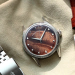 A silver-tone wristwatch with a natural burlwood dial and a fluted bezel, styled on a beige fabric surface next to a silver metal link bracelet and a brown leather strap.