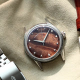 A silver-tone wristwatch with a natural burlwood dial and a fluted bezel, styled on a beige fabric surface next to a silver metal link bracelet and a brown leather strap.