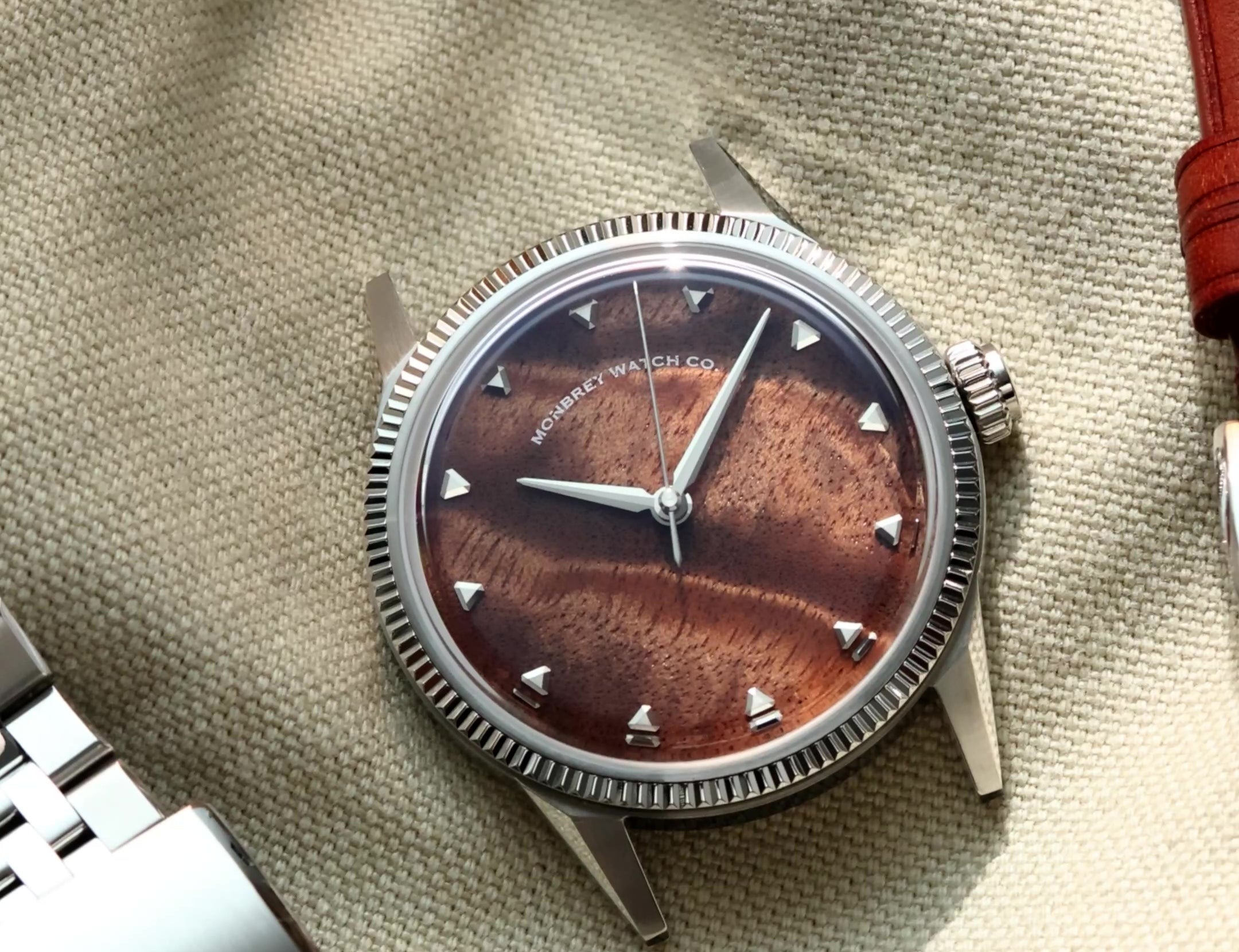 A silver-tone wristwatch with a natural burlwood dial and a fluted bezel, styled on a beige fabric surface next to a silver metal link bracelet and a brown leather strap.