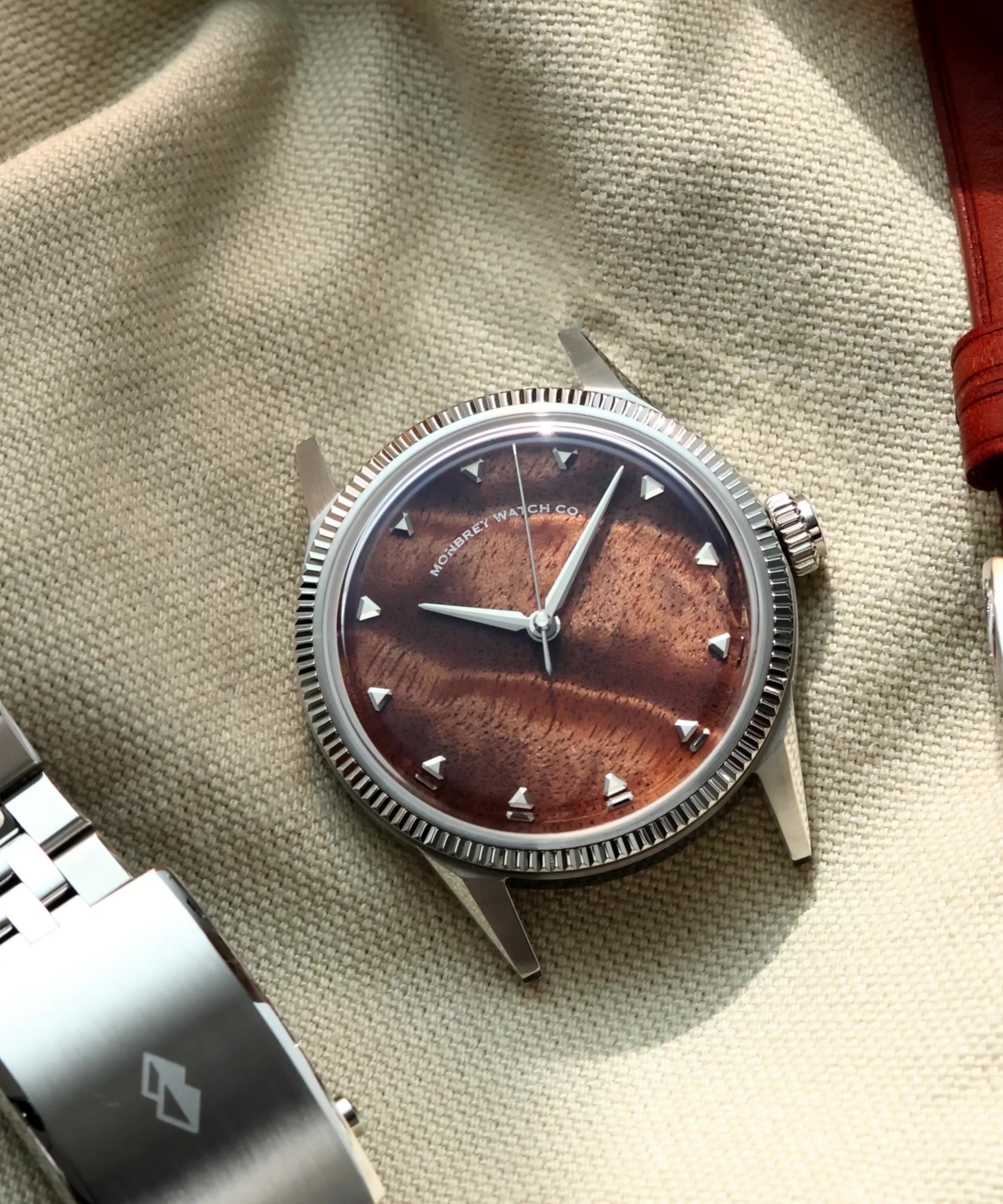 A silver-tone wristwatch with a natural burlwood dial and a fluted bezel, styled on a beige fabric surface next to a silver metal link bracelet and a brown leather strap.