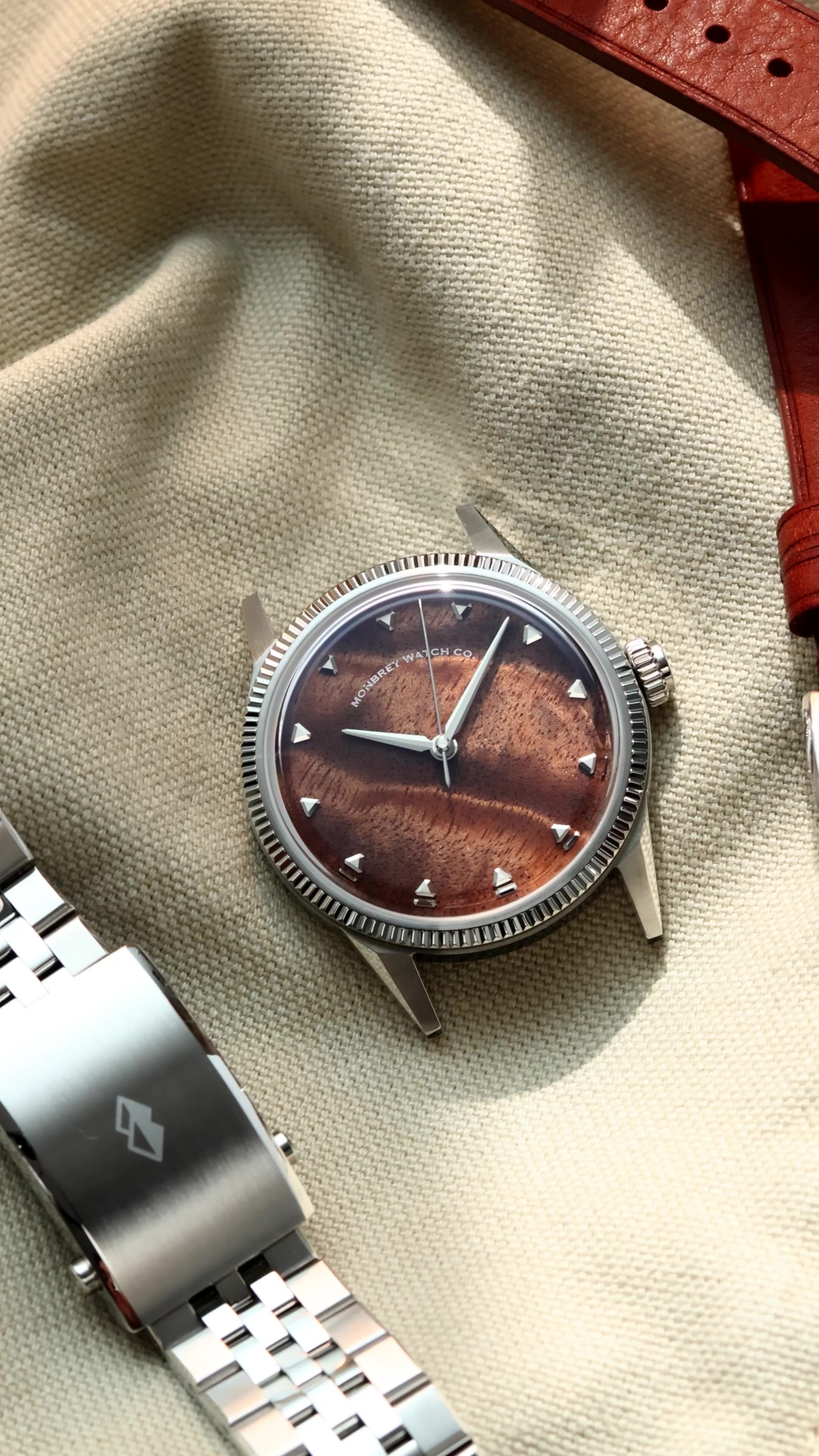 A silver-tone wristwatch with a natural burlwood dial and a fluted bezel, styled on a beige fabric surface next to a silver metal link bracelet and a brown leather strap.