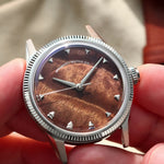 A close-up of a wristwatch being held in two hands. The watch has a silver-tone fluted bezel, a brown burlwood dial with a sunburst finish, and silver-tone hour markers.