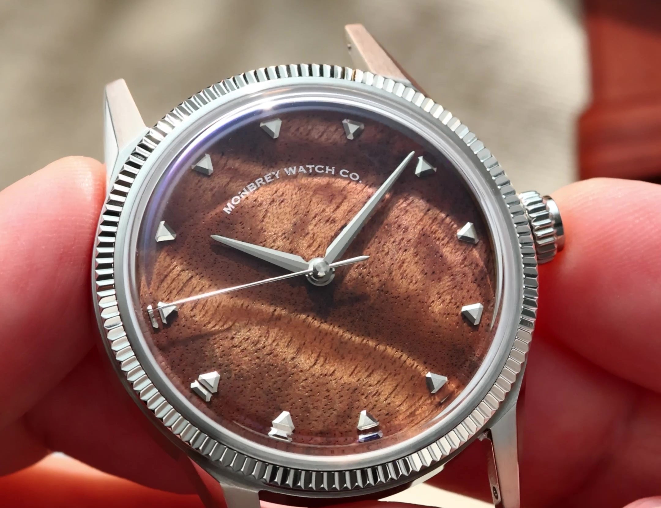 A close-up of a wristwatch being held in two hands. The watch has a silver-tone fluted bezel, a brown burlwood dial with a sunburst finish, and silver-tone hour markers.