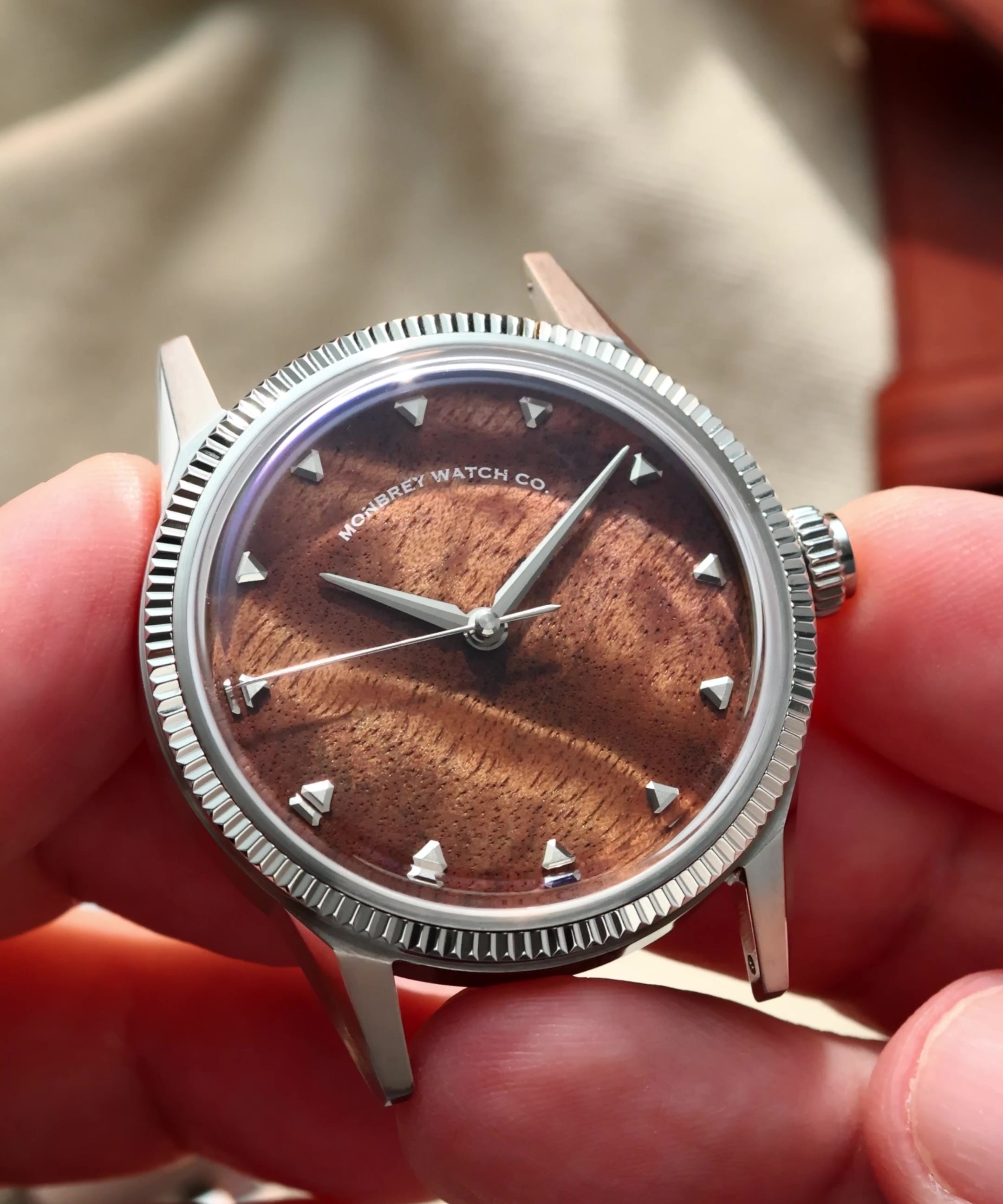 A close-up of a wristwatch being held in two hands. The watch has a silver-tone fluted bezel, a brown burlwood dial with a sunburst finish, and silver-tone hour markers.