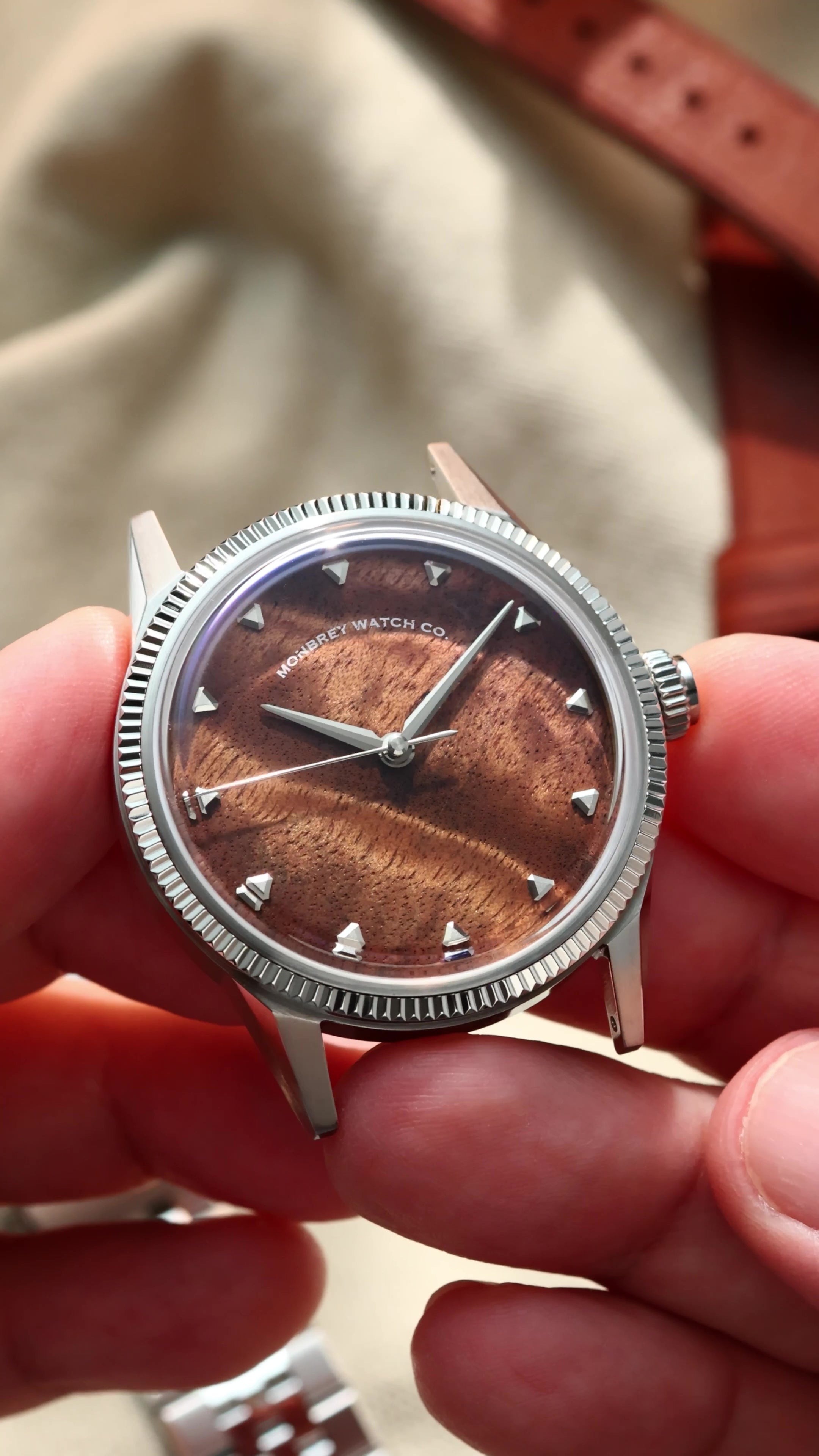 A close-up of a wristwatch being held in two hands. The watch has a silver-tone fluted bezel, a brown burlwood dial with a sunburst finish, and silver-tone hour markers.